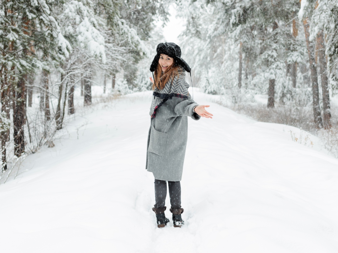 Девушка в пальто и теплой шапке гуляет в заснеженном лесу 