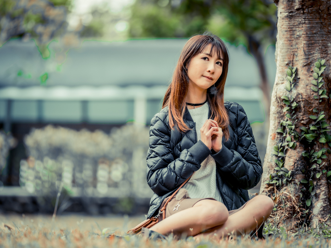 Девушка азиатка в черной куртке сидит под деревом