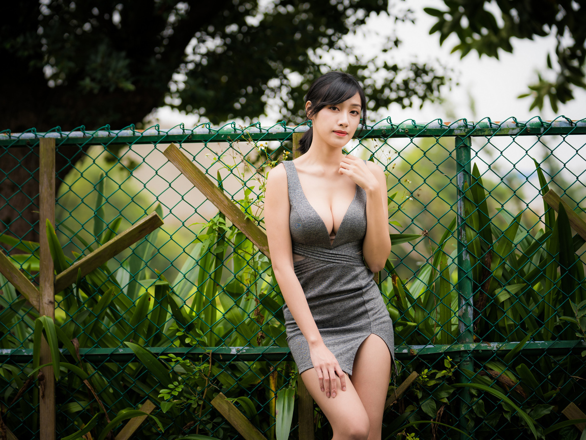 Красивая азиатка в сером платье стоит у зеленой сетки 