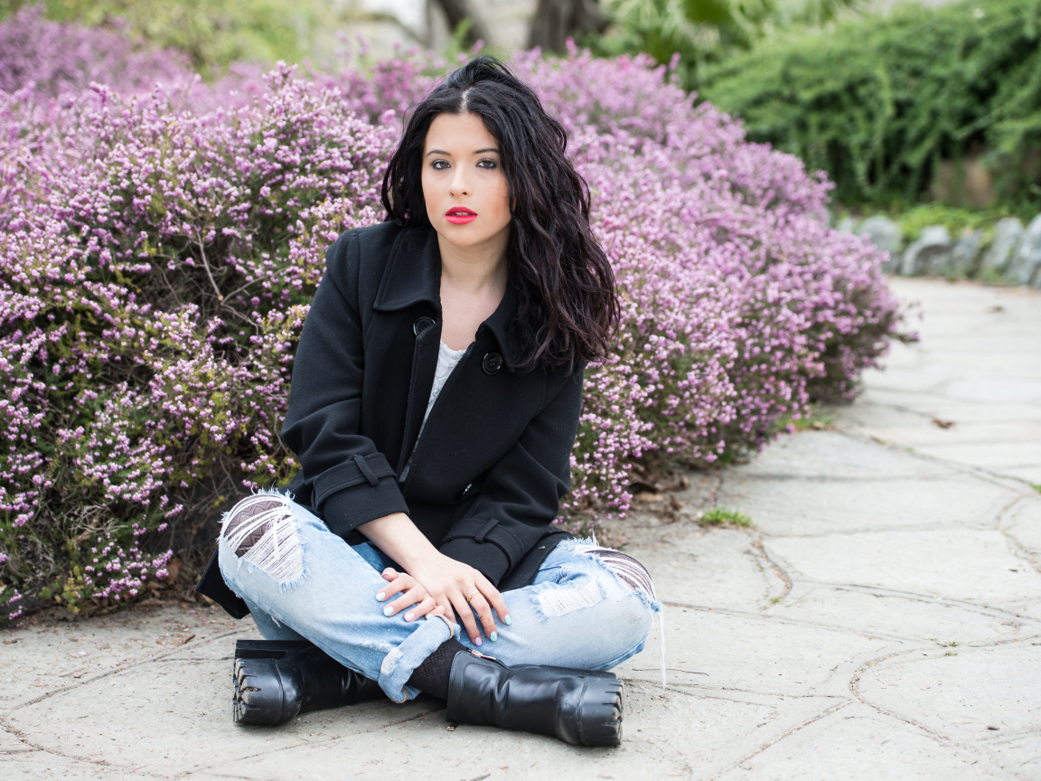 Красивая девушка в черном пальто сидит на земле у кустов