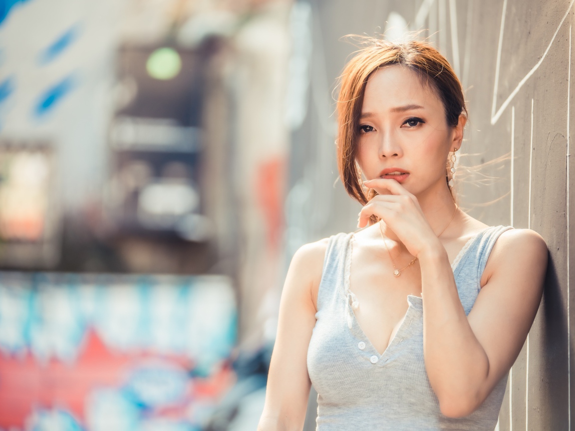 Милая девушка азиатка  с рукой у лица стоит у стены 
