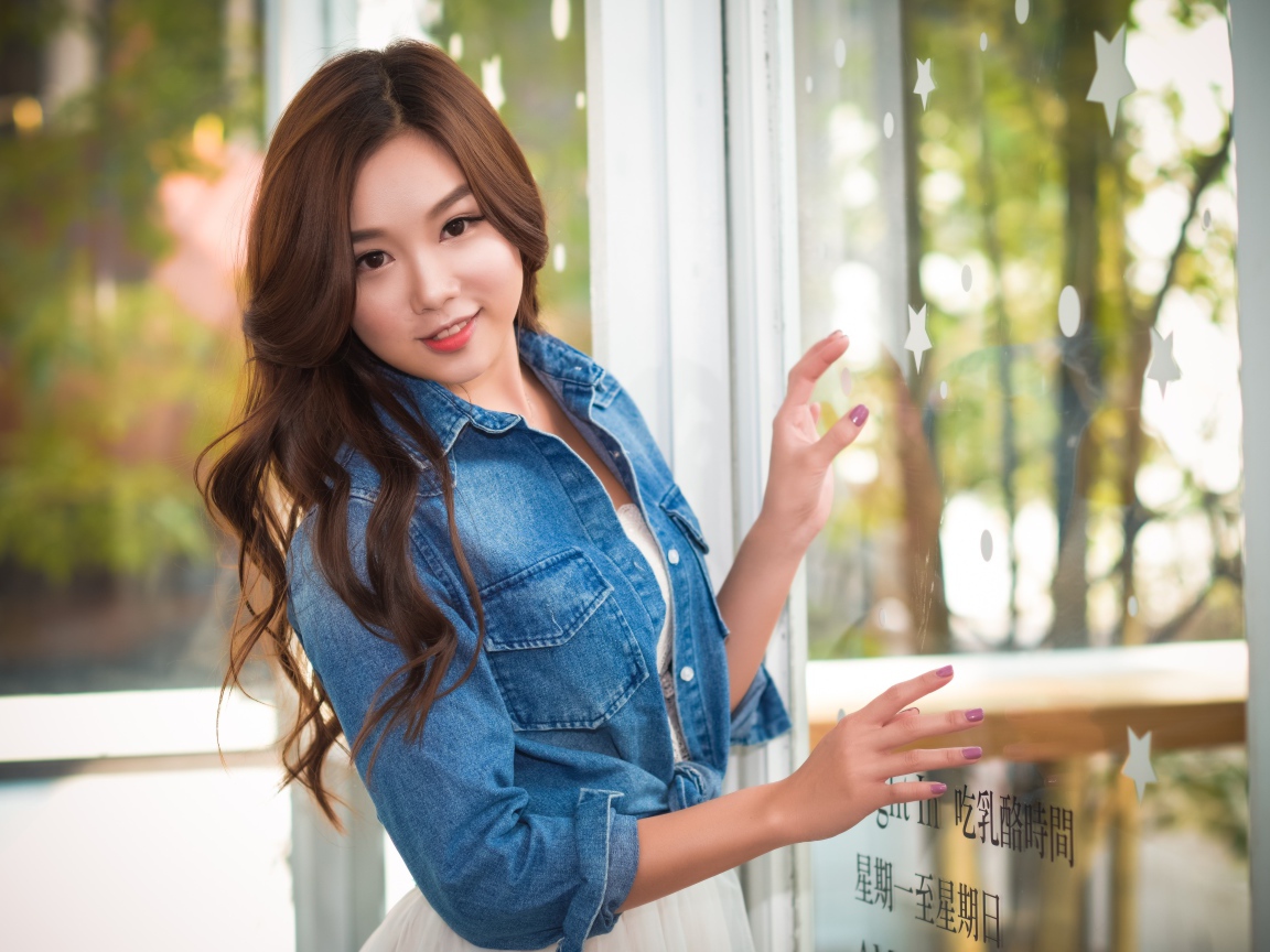 Милая девушка азиатка в джинсовой куртке стоит у окна 