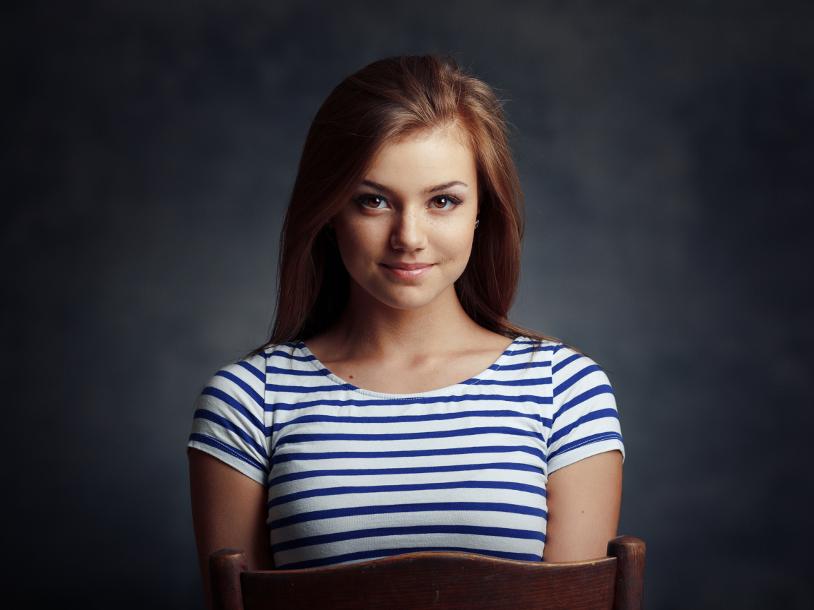 Милая кареглазая девушка в полосатой майке сидит на стуле 