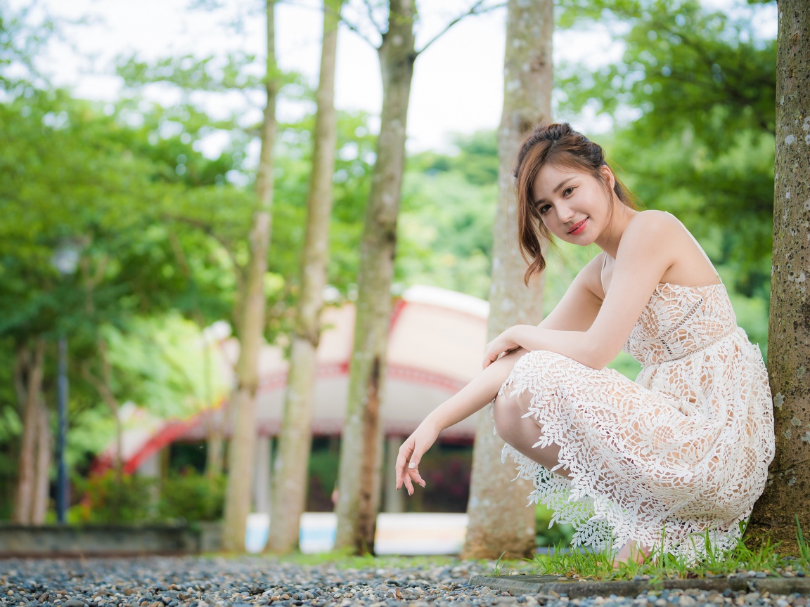 Улыбающаяся девушка в кружевном платье сидит под деревом в парке