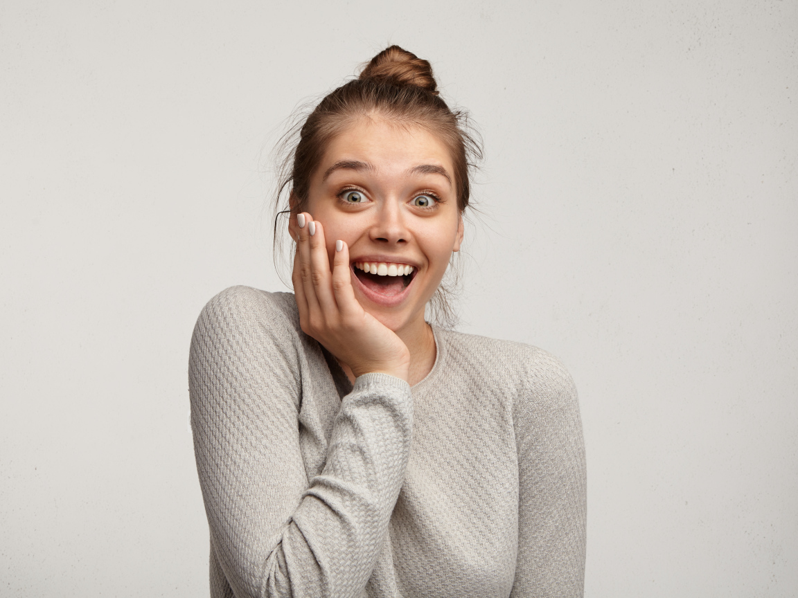 Удивленная девушка с рукой у лица на сером фоне 