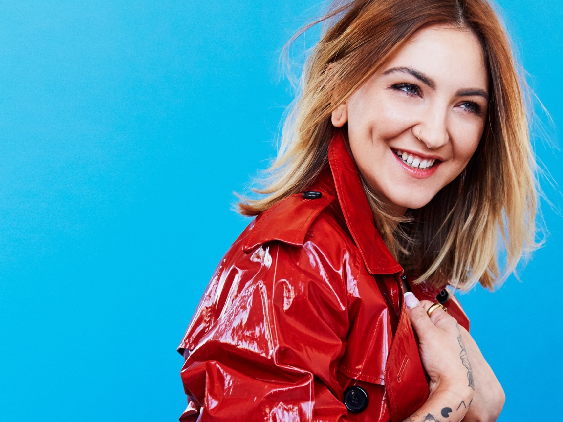 Улыбающаяся певица Джулия Майклз в красной куртке на голубом фоне