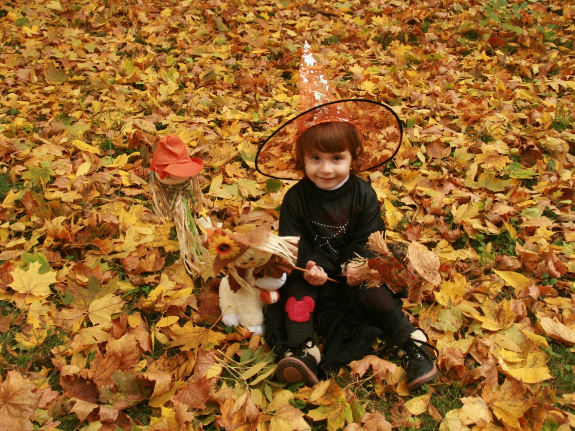Маленькая девочка в костюме ведьмы сидит на опавшей листве на Хэллоуин