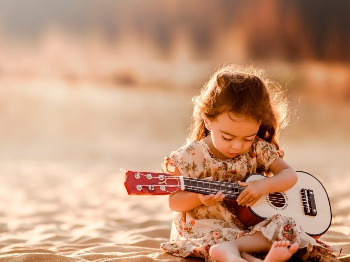 Маленькая девочка с гитарой сидит на песке 