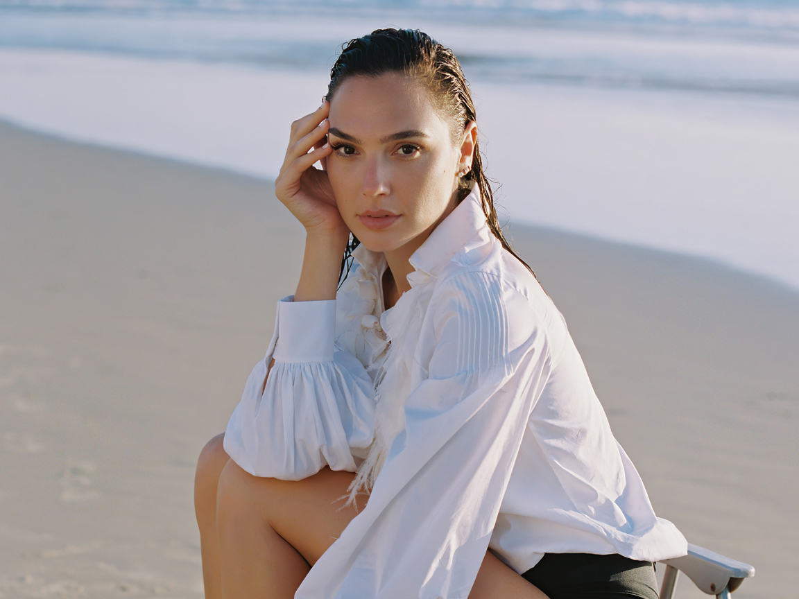 Актриса Галь Гадот в белой рубашке сидит на песке