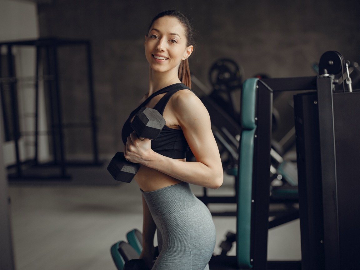 Улыбающаяся спортивная стройная девушка в спортзале с гантелями