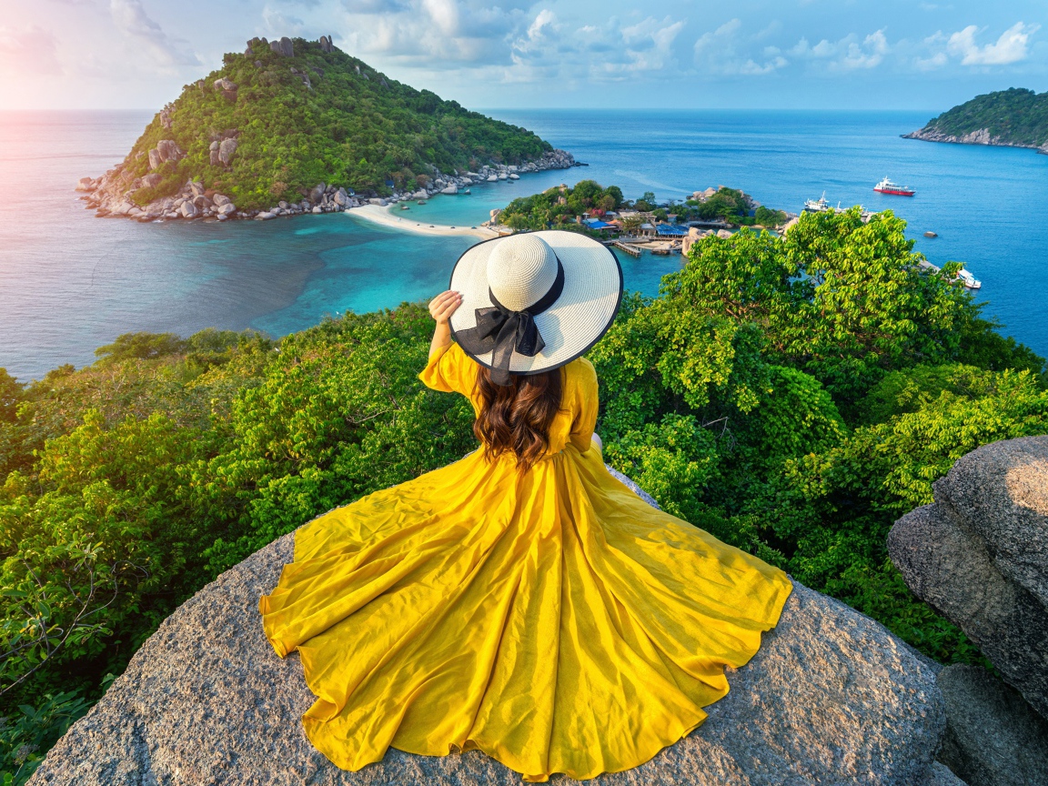 A girl in a beautiful yellow dress sits on a stone overlooking the sea