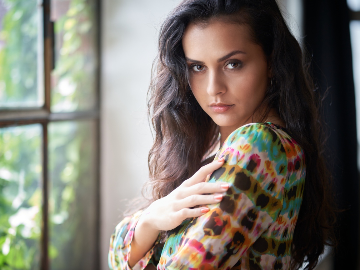 Длинноволосая кареглазая девушка стоит у окна