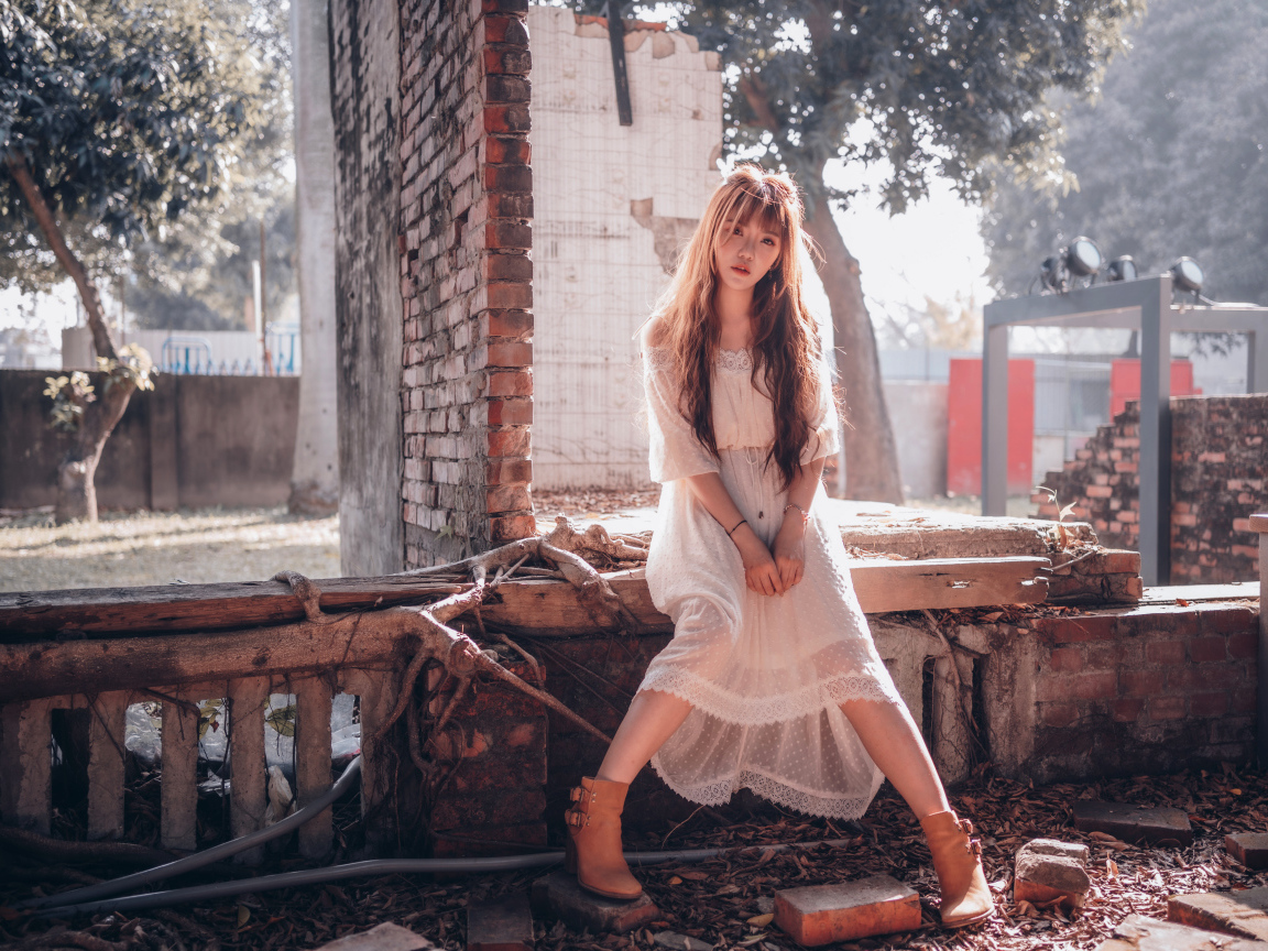 Молодая девушка азиатка в белом платье сидит на разваленной стене