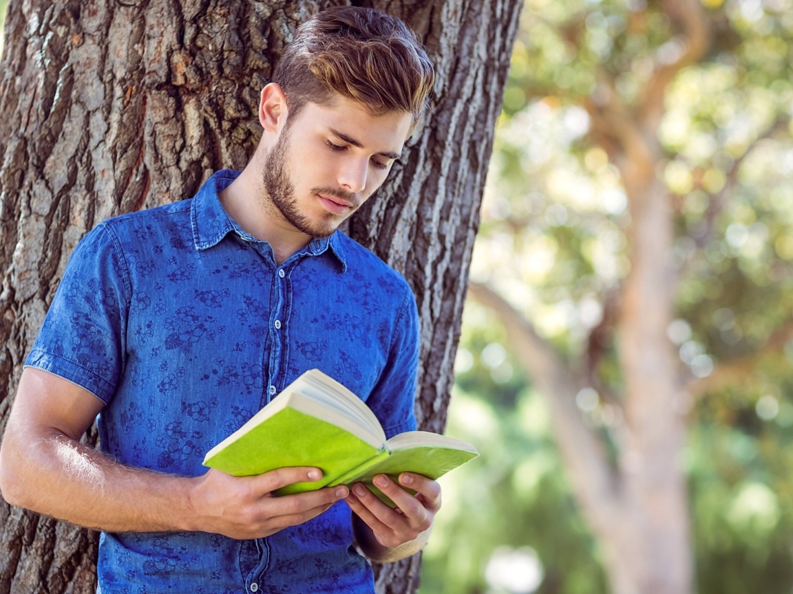 Красивый молодой мужчина стоит с книгой под деревом 