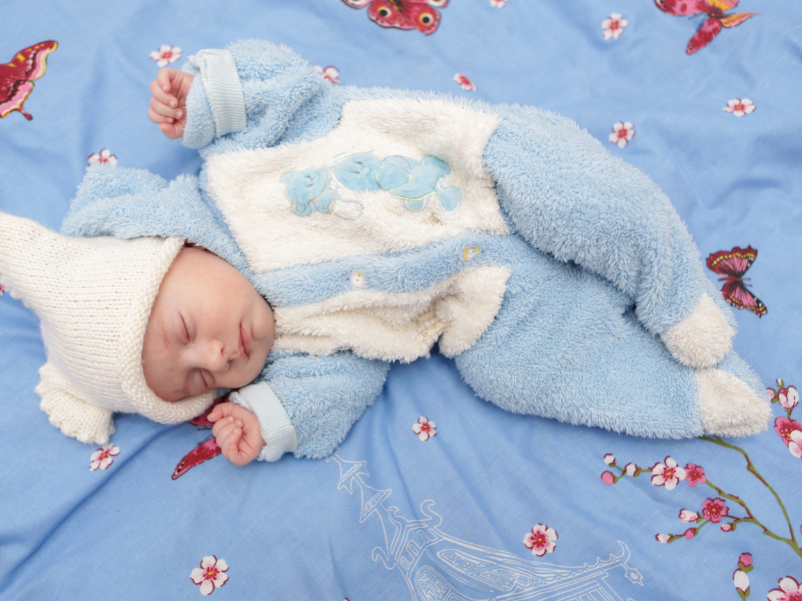 Грудной ребенок в голубом костюме лежит на кровати 