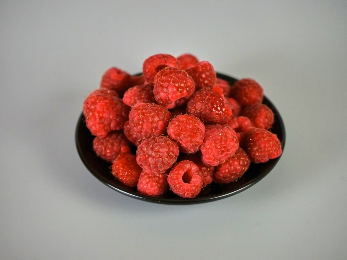 Спелая красная малина в черной тарелке на сером фоне