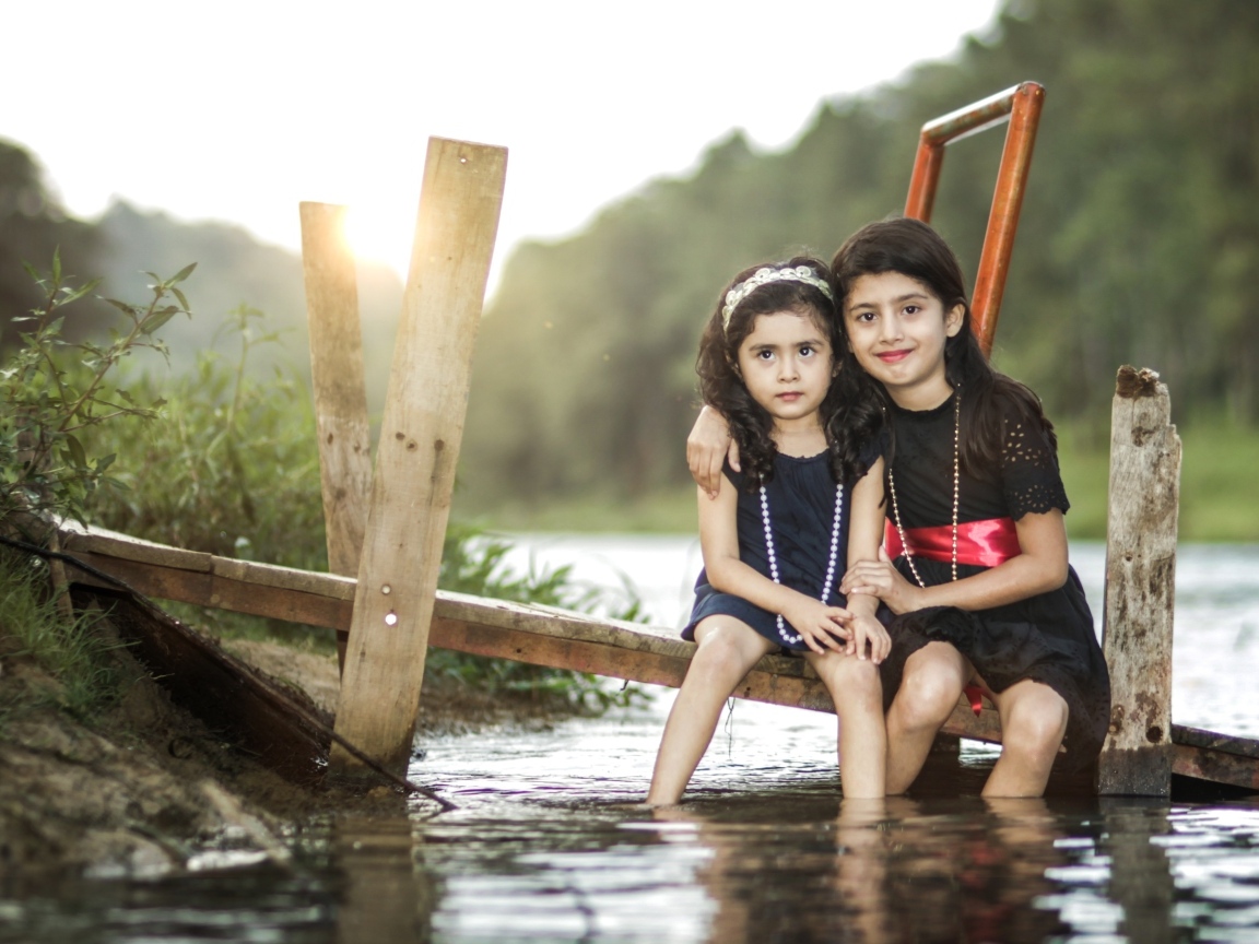 Две девочки сидят на мосту у реки