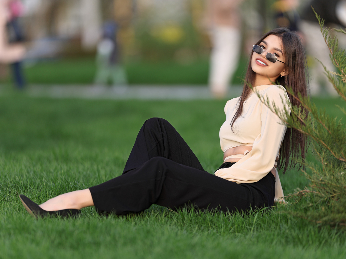 Девушка в черных брюках и блузке сидит на траве