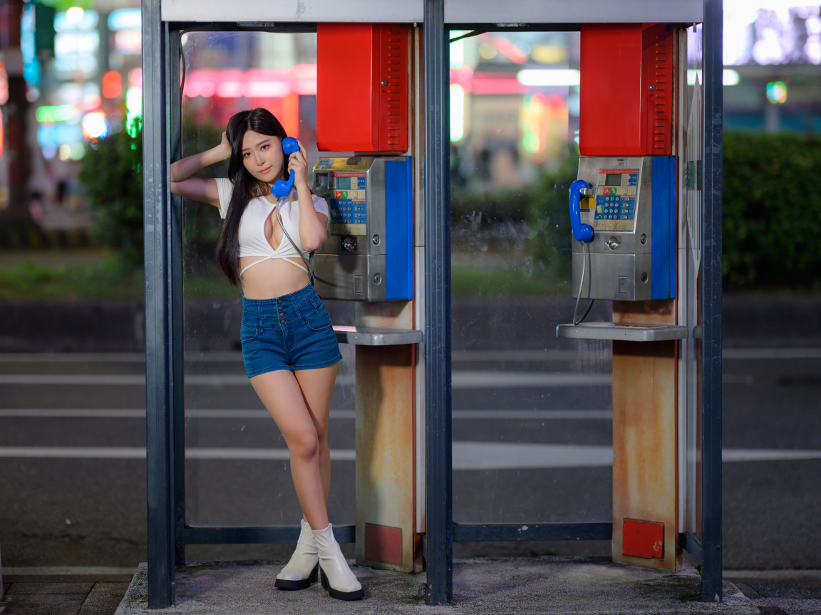 Азиатка в коротких шортах стоит у телефона-автомата
