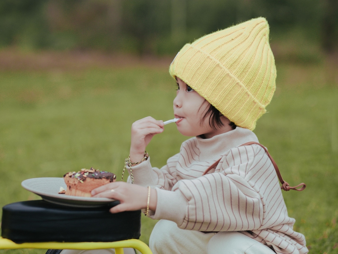 Маленькая девочка в желтой шапке ест пирожное