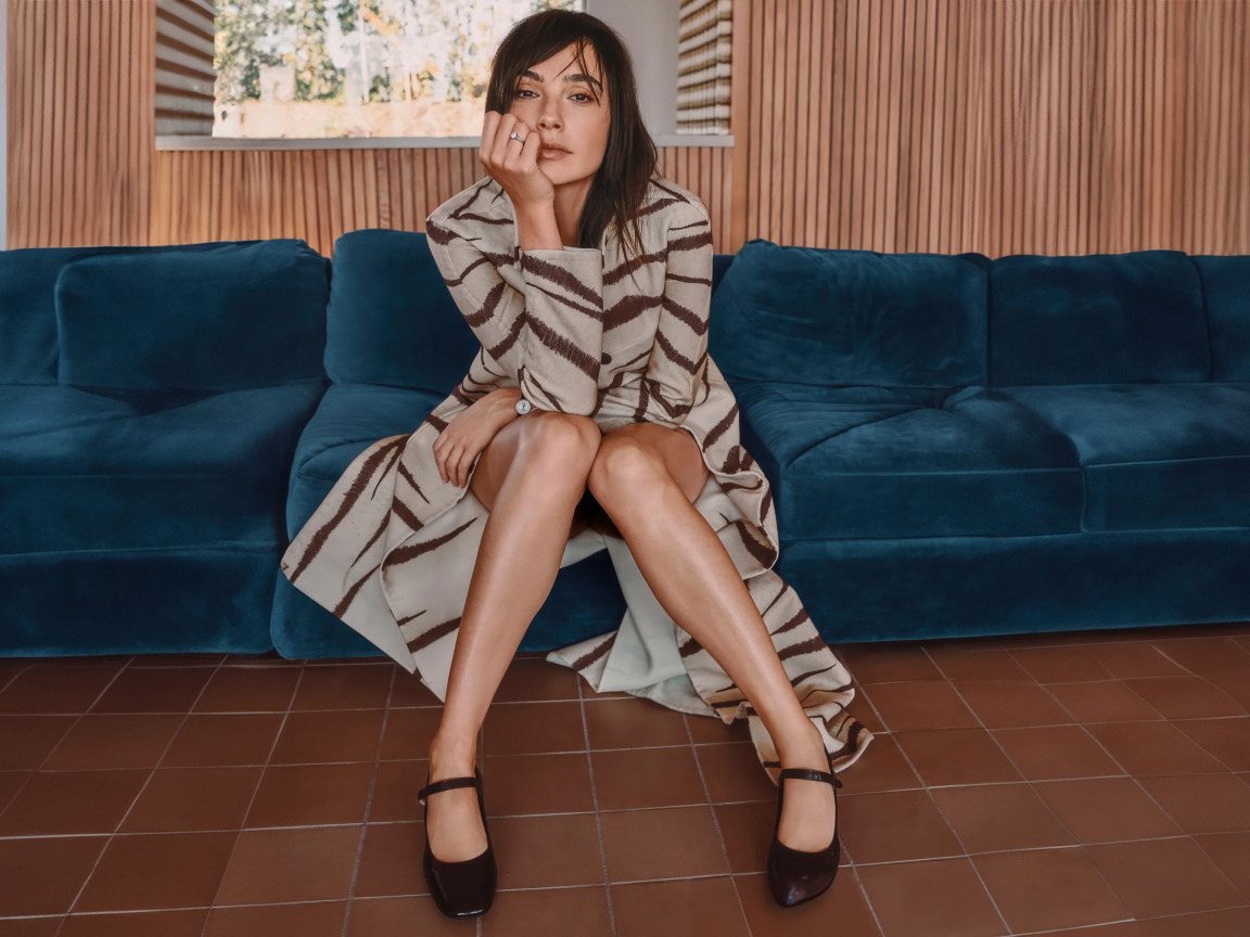 Актриса Галь Гадот в пальто сидит на диване