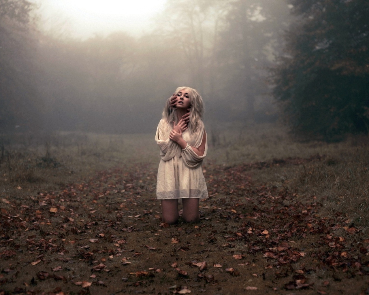 Блондинка в белом платье стоит на коленях среди леса