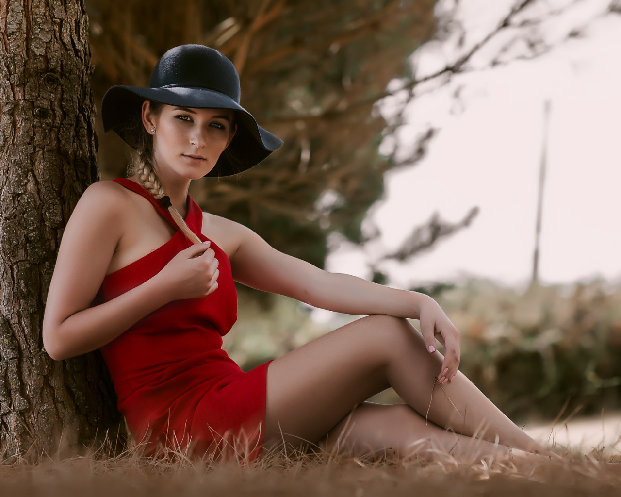 Красивая девушка в красном платье и черной шляпе сидит под деревом