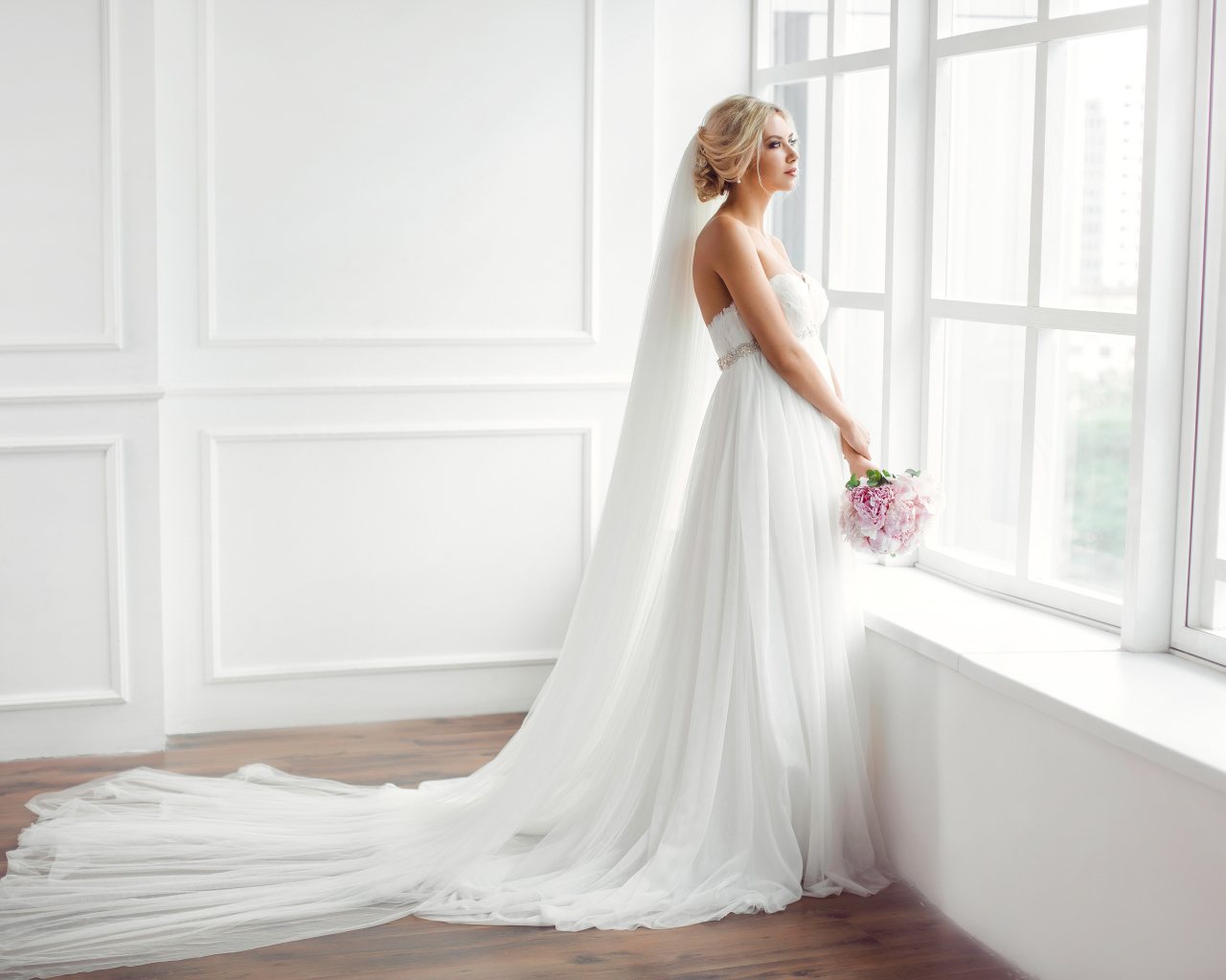 Красивая девушка в белом свадебном платье с букетом стоит у окна