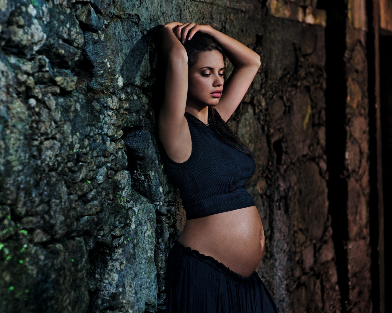 Беременная девушка в черном наряде у стены 