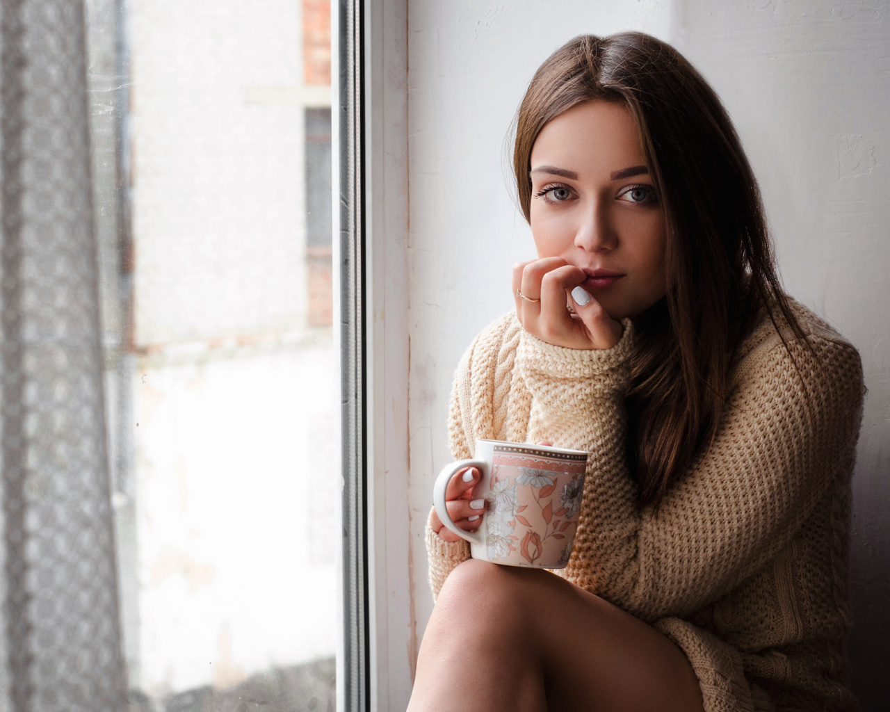 Молодая девушка в свитере сидит с чашкой на подоконнике