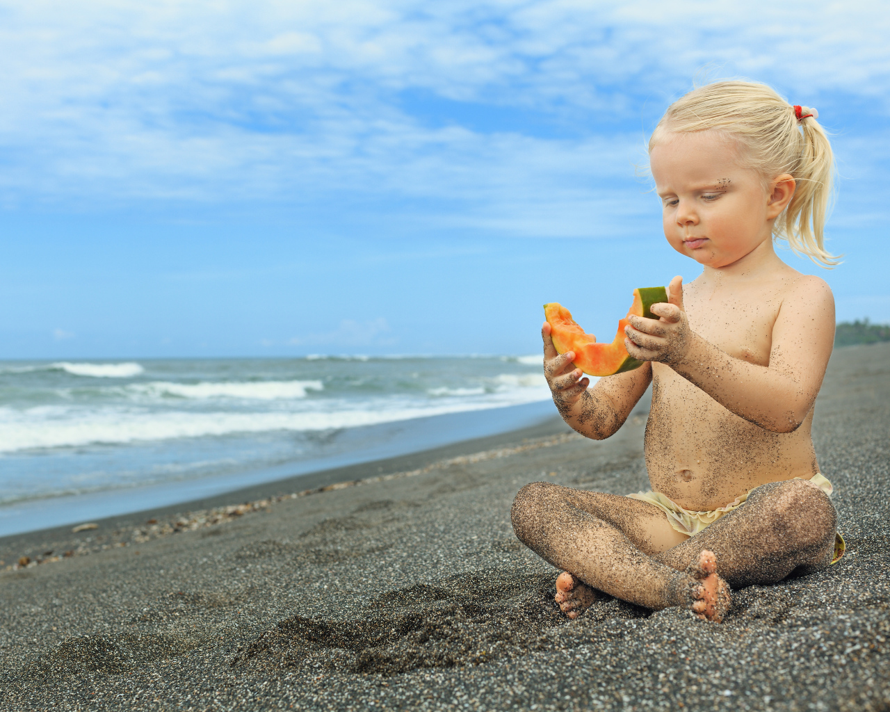 Маленькая девочка сидит на песке у моря с кусочком дыни