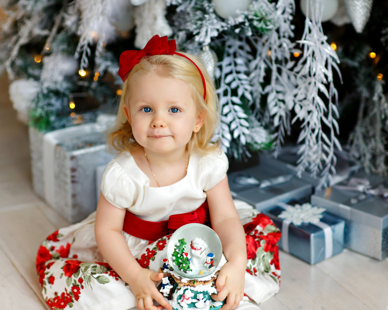 Маленькая девочка блондинка сидит с подарками под новогодней елкой