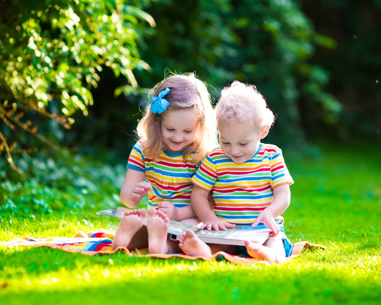 Маленькие мальчик и девочка читают книгу сидя на зеленой траве