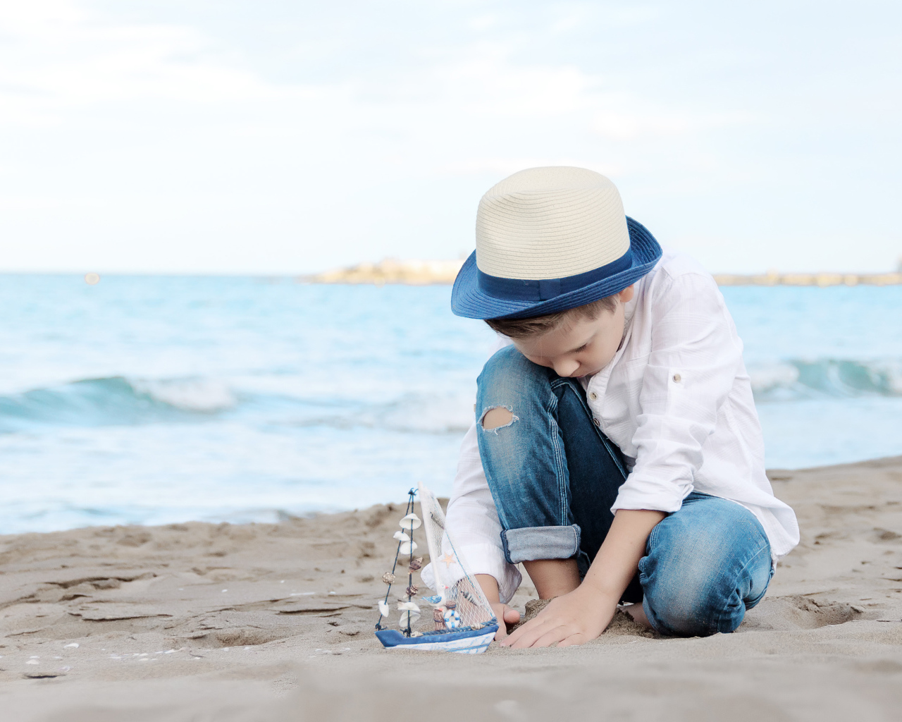 Маленький мальчик в шляпе играет с игрушечным корабликом на песке