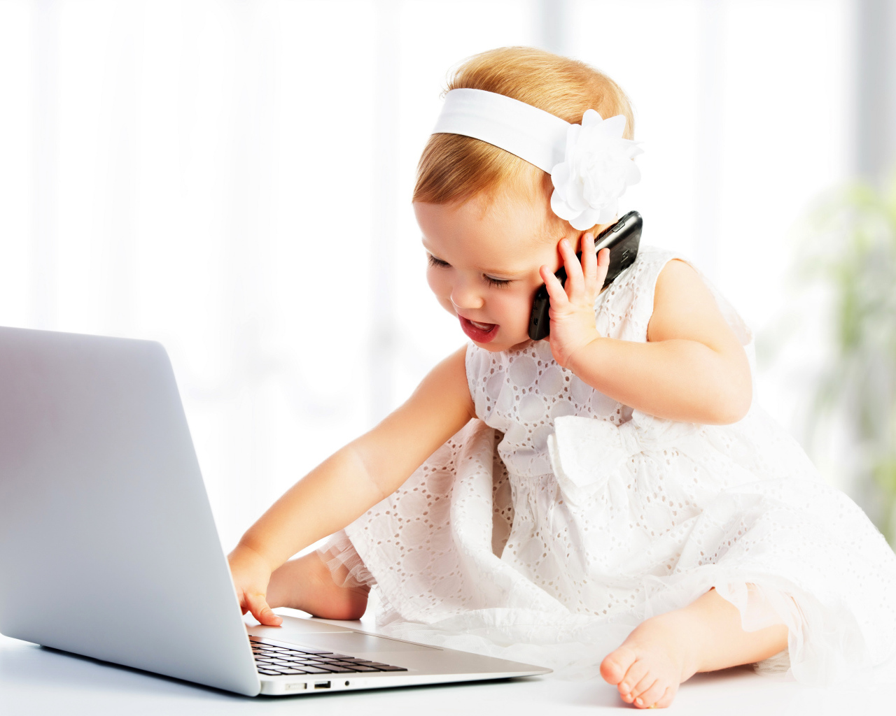 Маленькая девочка в белом платье сидит с телефоном у ноутбука