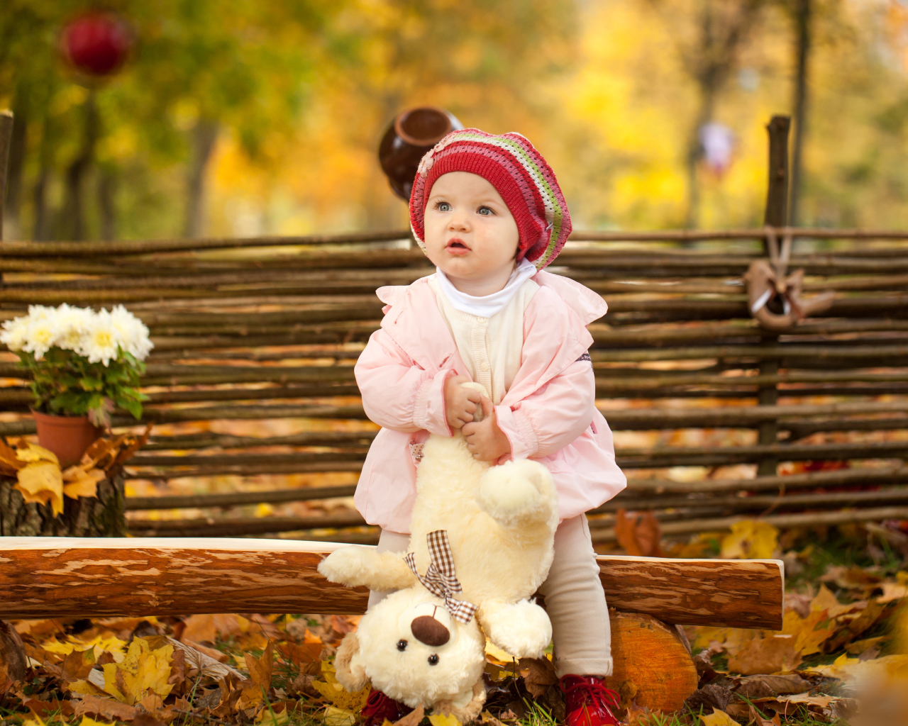 Маленькая девочка с плюшевым мишкой сидит на деревянной лавке осенью