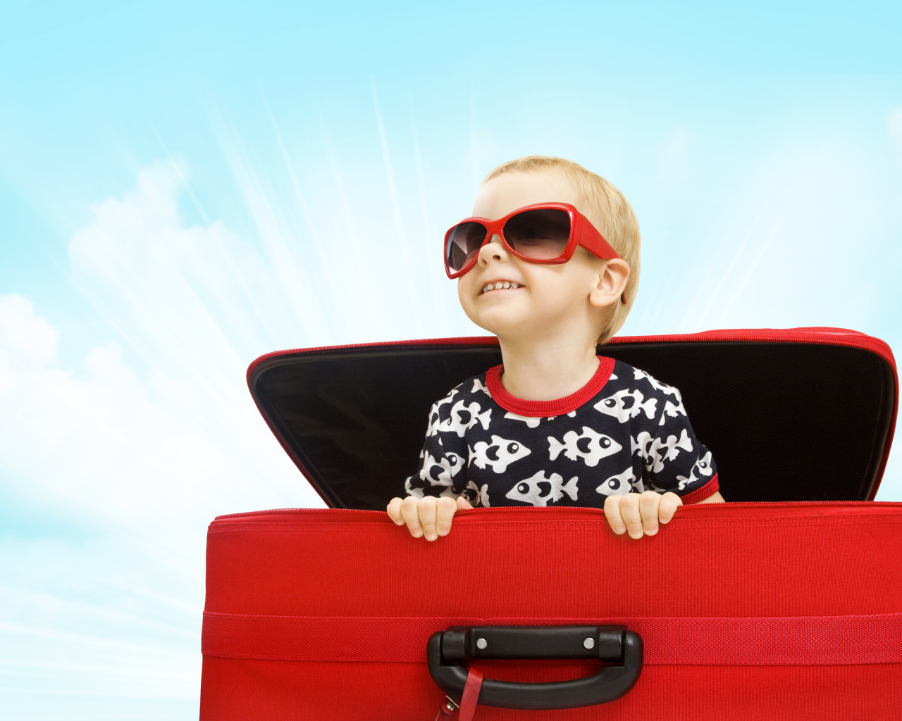 Маленький улыбающийся мальчик в очках сидит в красном чемодане