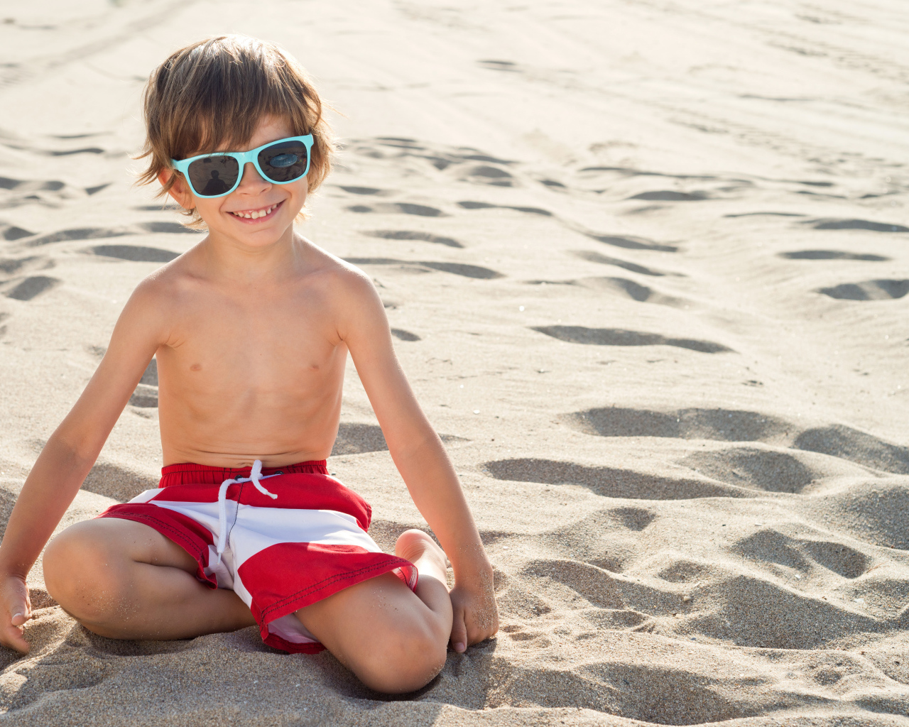 Маленький улыбающийся мальчик в очках сидит на песке