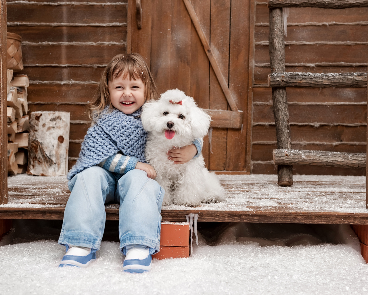 Маленькая улыбающаяся девочка сидит с белым пуделем на крыльце дома зимой