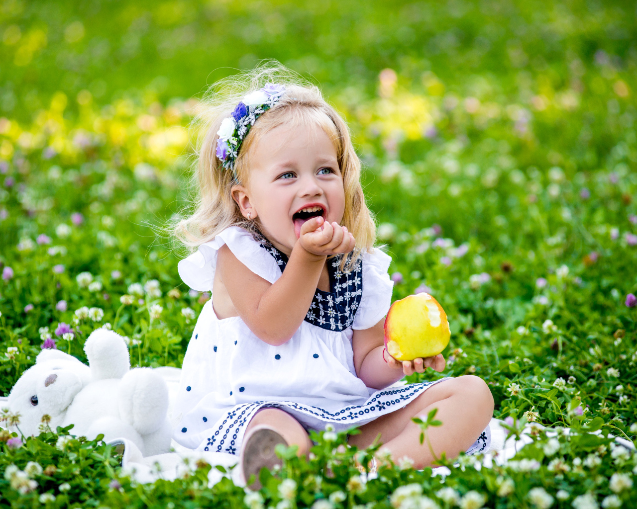 Маленькая улыбающаяся девочка с яблоком сидит на зеленой траве