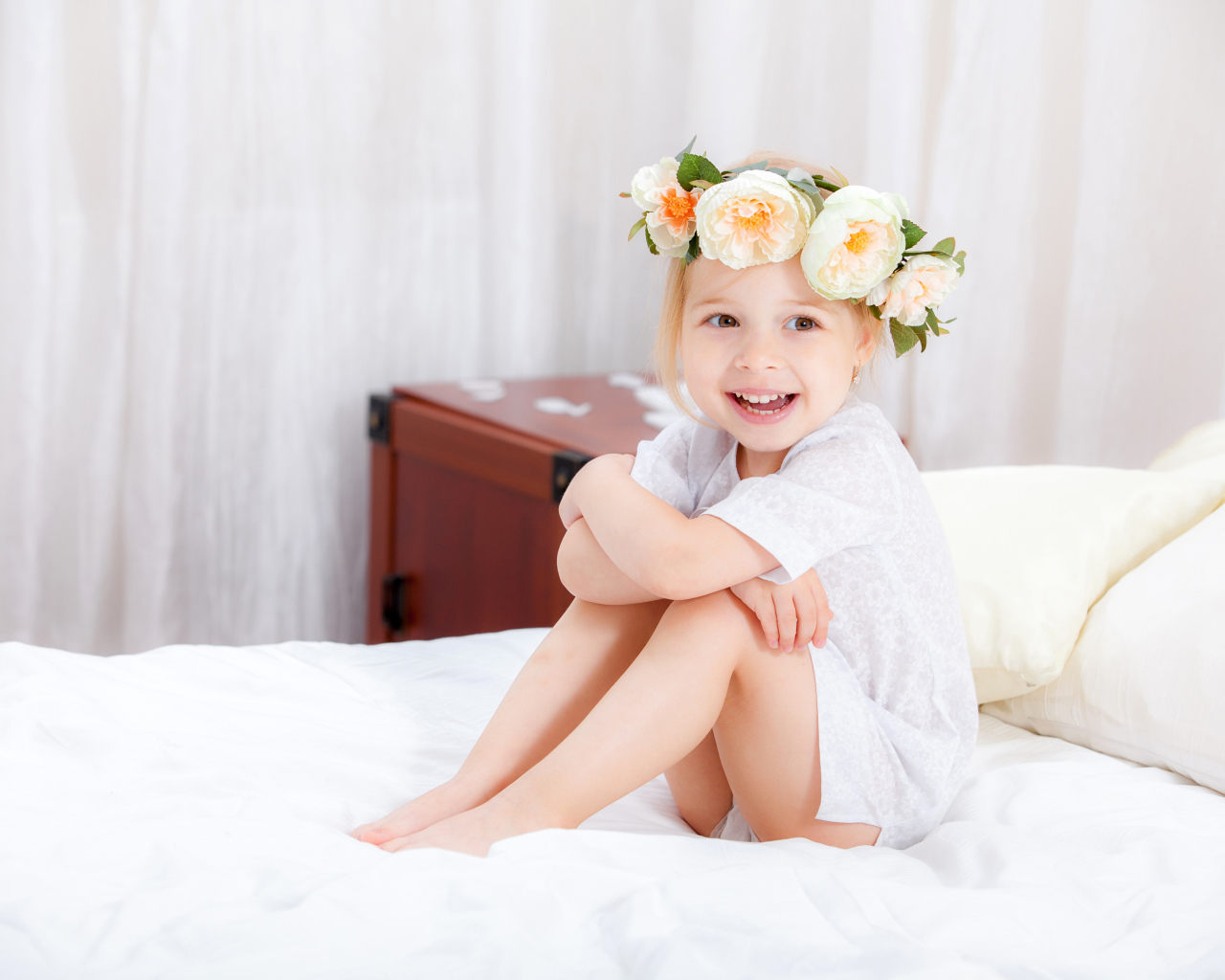 Улыбающаяся девочка с венком на голове сидит на кровати