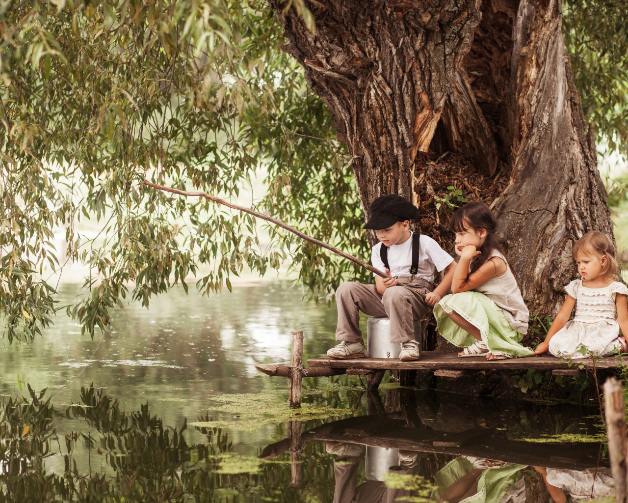 Две маленькие девочки и мальчик сидят на берегу пруда с удочкой