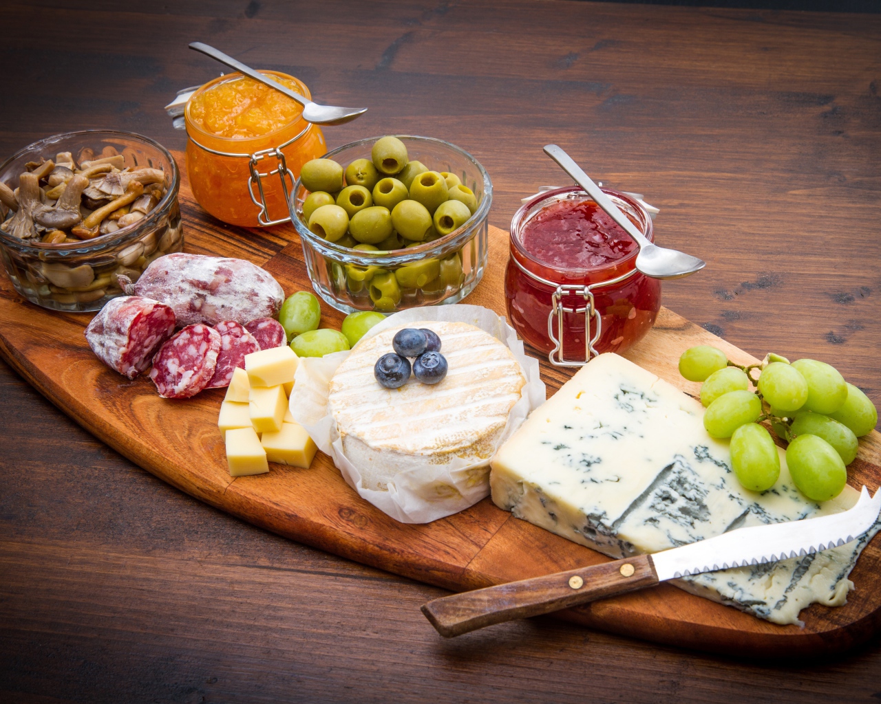 Сыр, грибы, маслины, колбаса, виноград и джем на разделочной доске 
