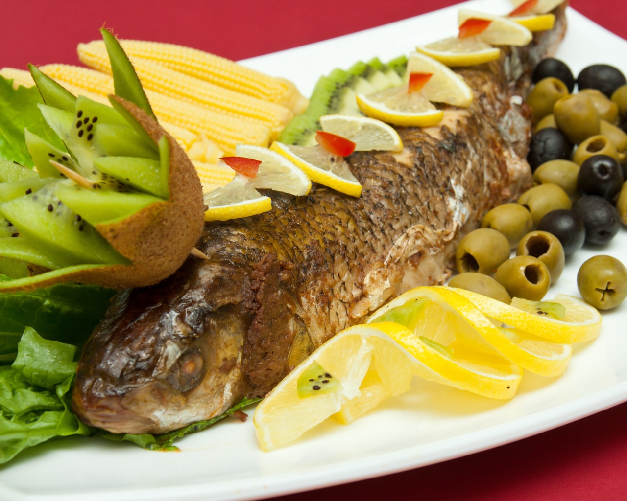 Запеченная рыба с оливками, лимоном, киви, кукурузой и листьями салата на белой тарелке