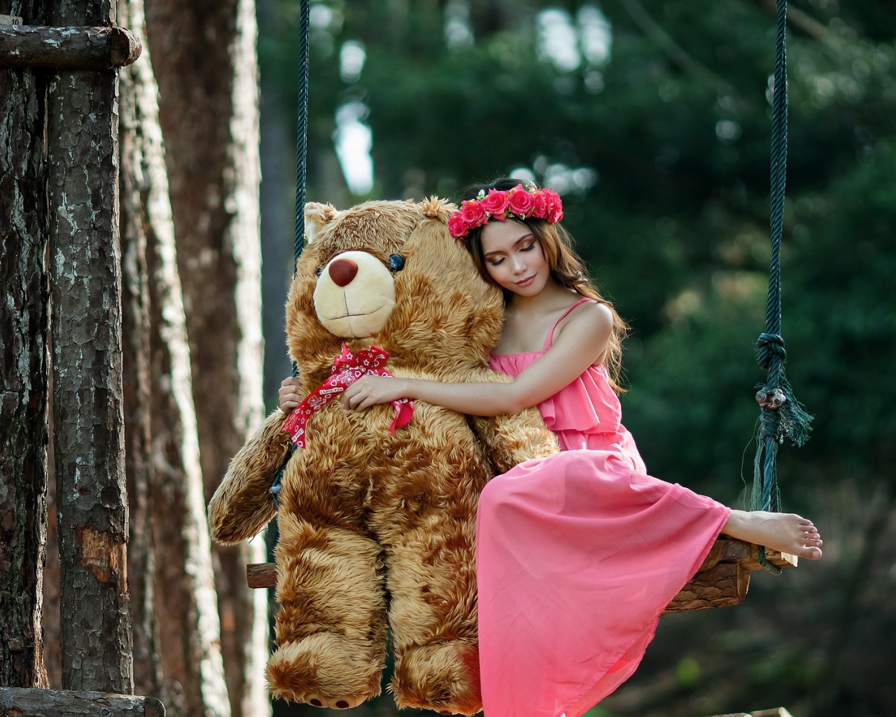 Красивая девушка азиатка в розовом платье сидит на качели с большим игрушечным медведем
