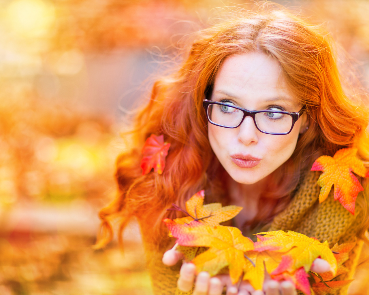 Рыжеволосая девушка в очках держит в руках опавшие листья 