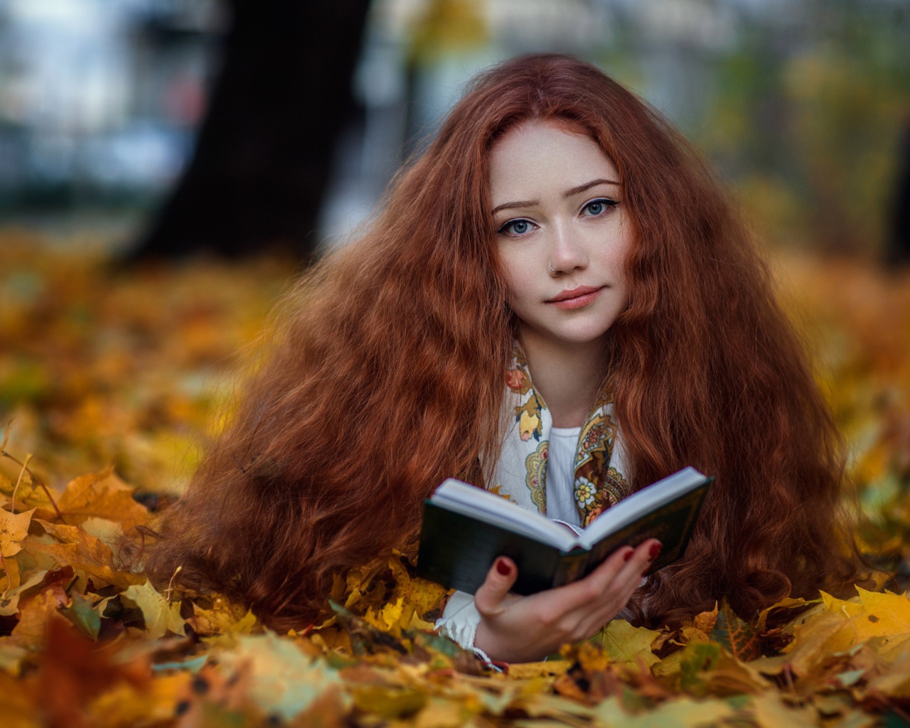 Рыжеволосая девушка с книгой лежит на опавших листьях осенью 
