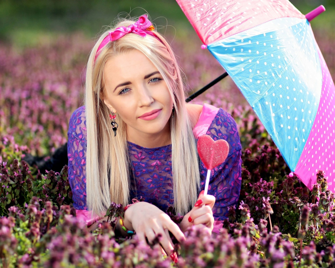 Молодая девушка блондинка лежит на поляне с сиреневыми цветами под разноцветным зонтом