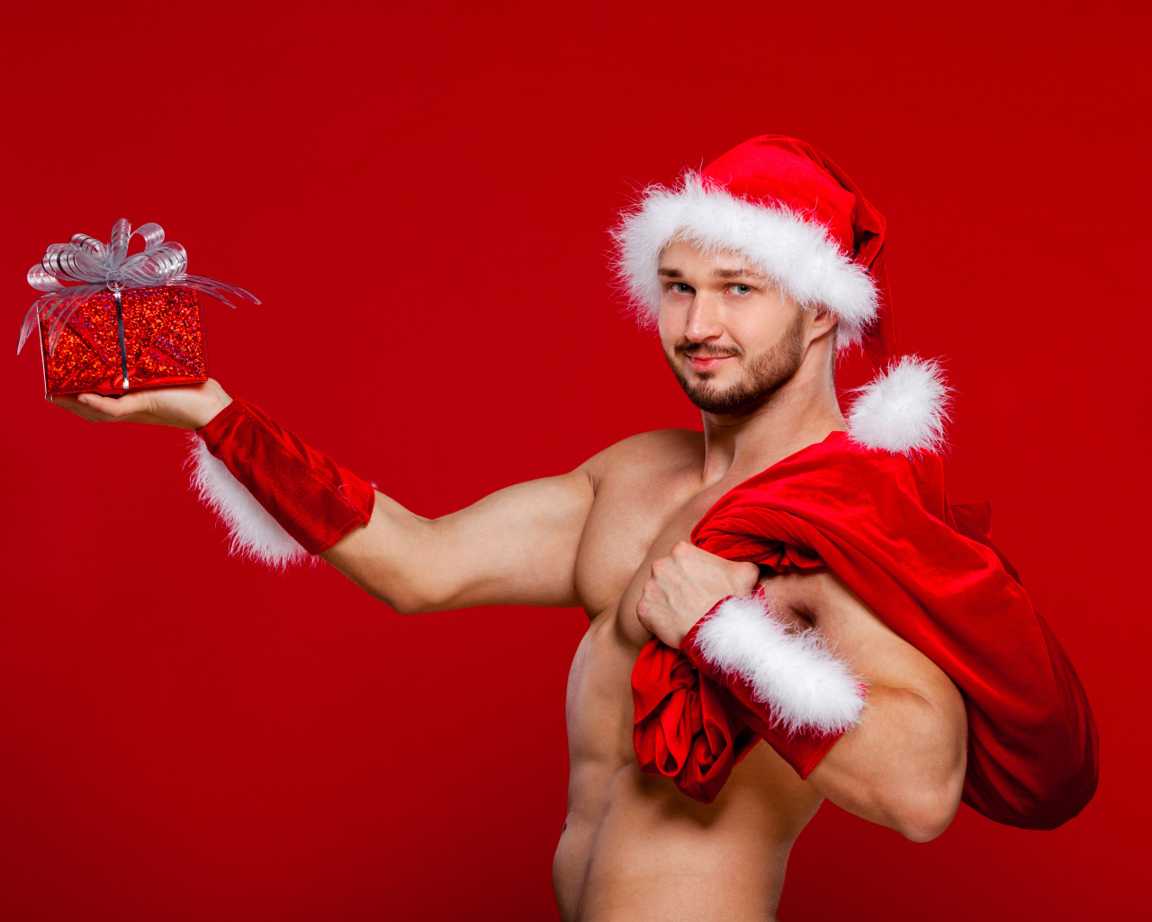 Мужчина в новогоднем костюме Санта Клауса с мешком и подарком в руке на красном фоне