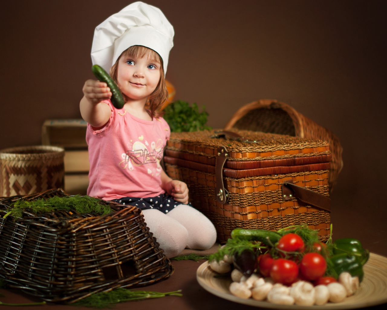 Маленькая девочка в шапке повара с огурцом в руках
